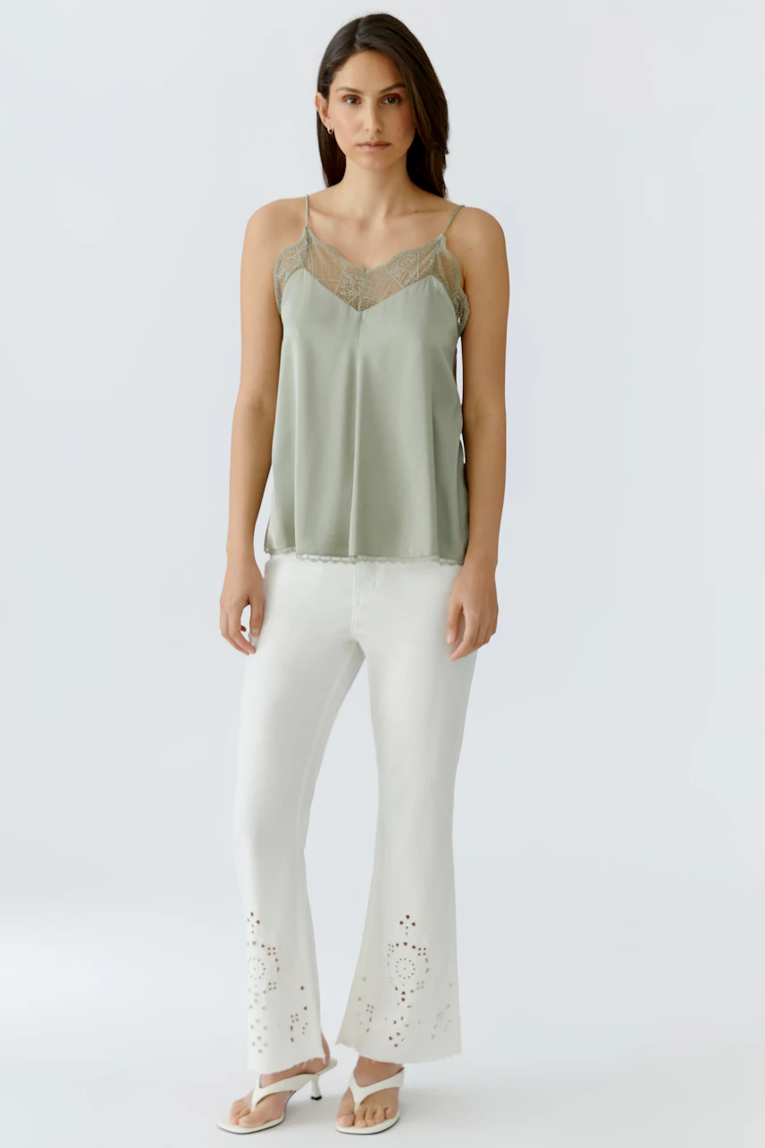Casual women's top: Woman in a green lace-trimmed top with white pants and sandals, standing confidently.