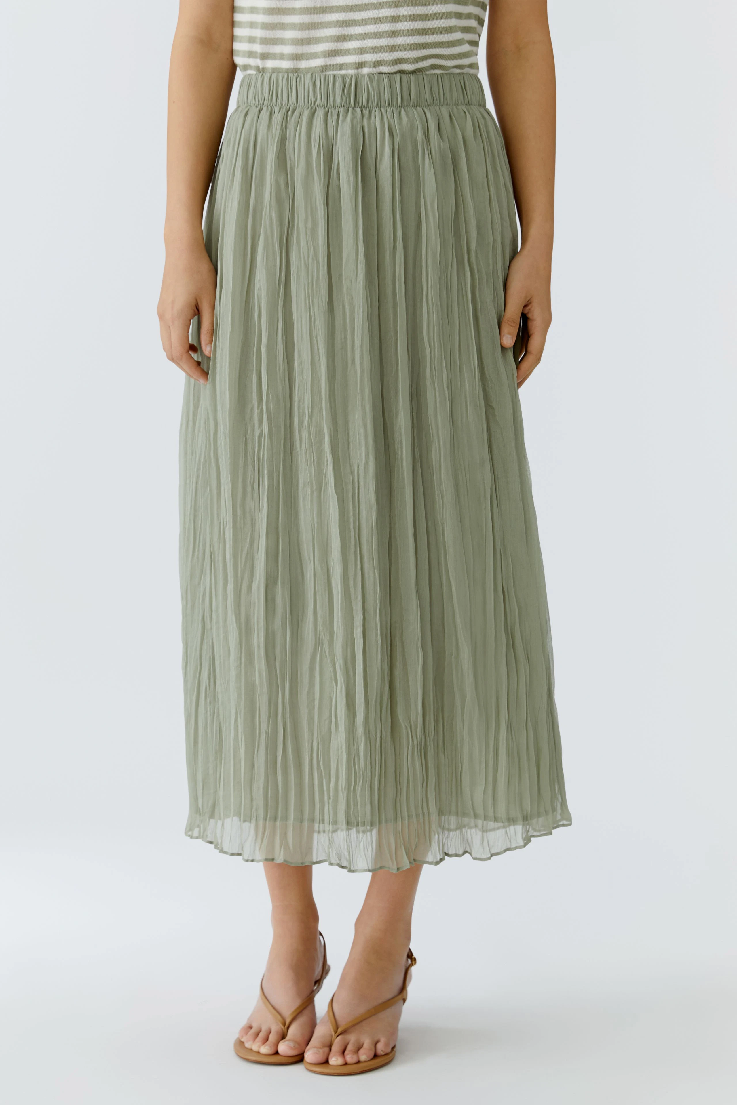Woman in a green pleated skirt, paired with a striped top, standing in sandals.