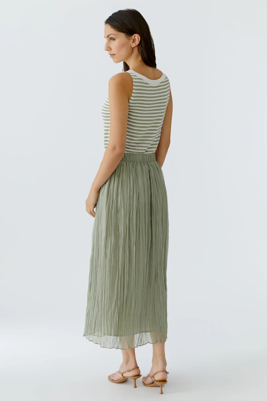 Woman in a striped tank top and green pleated skirt, standing sideways, showcasing casual style.