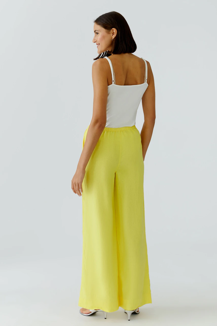 Woman in yellow Marlene pants and white top, standing confidently. The pants are wide-legged and flowy.