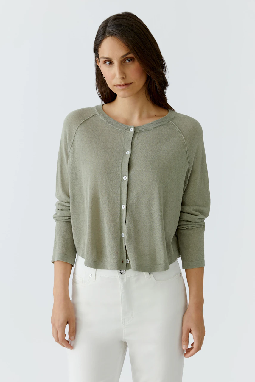 Damen-Strickjacke: Frau in olivgrüner Strickjacke mit Knopfleiste und weißen Hosen schaut direkt.