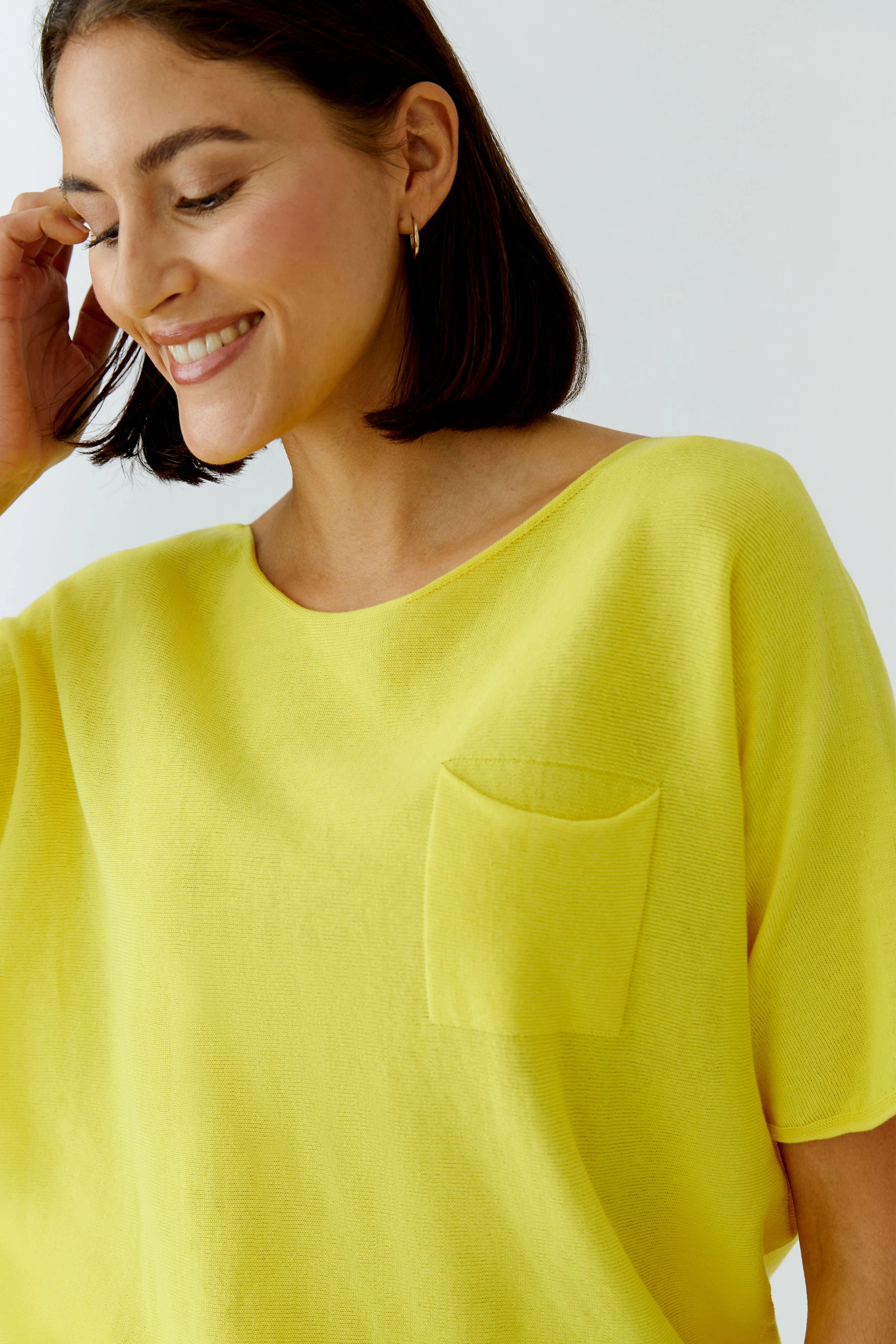Frau im gelben Pullover mit Brusttasche, lächelt und berührt ihr Haar. Schlichtes, modernes Design.