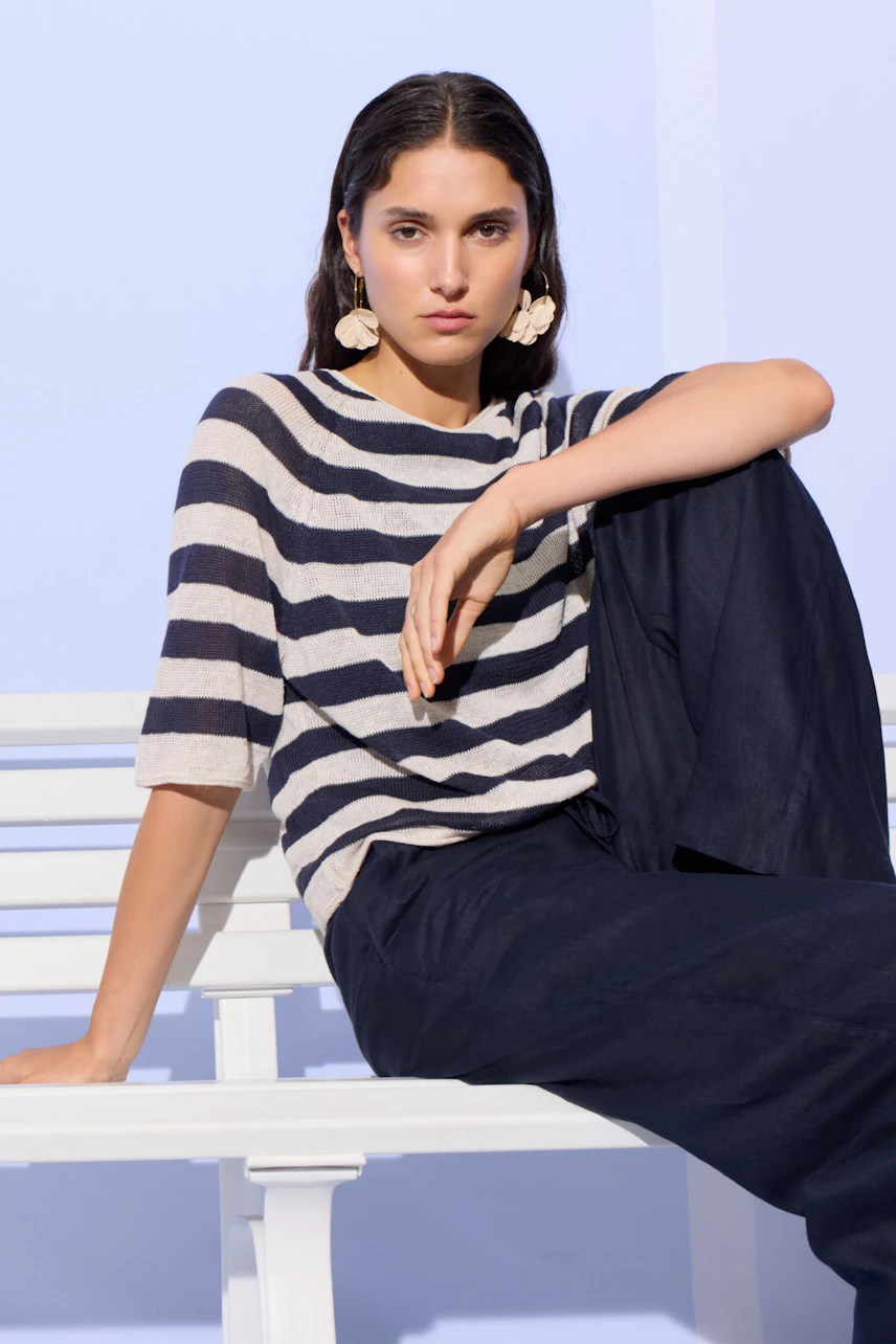 Casual women's pullover: Woman in striped pullover and dark pants sitting on a white bench, looking relaxed.