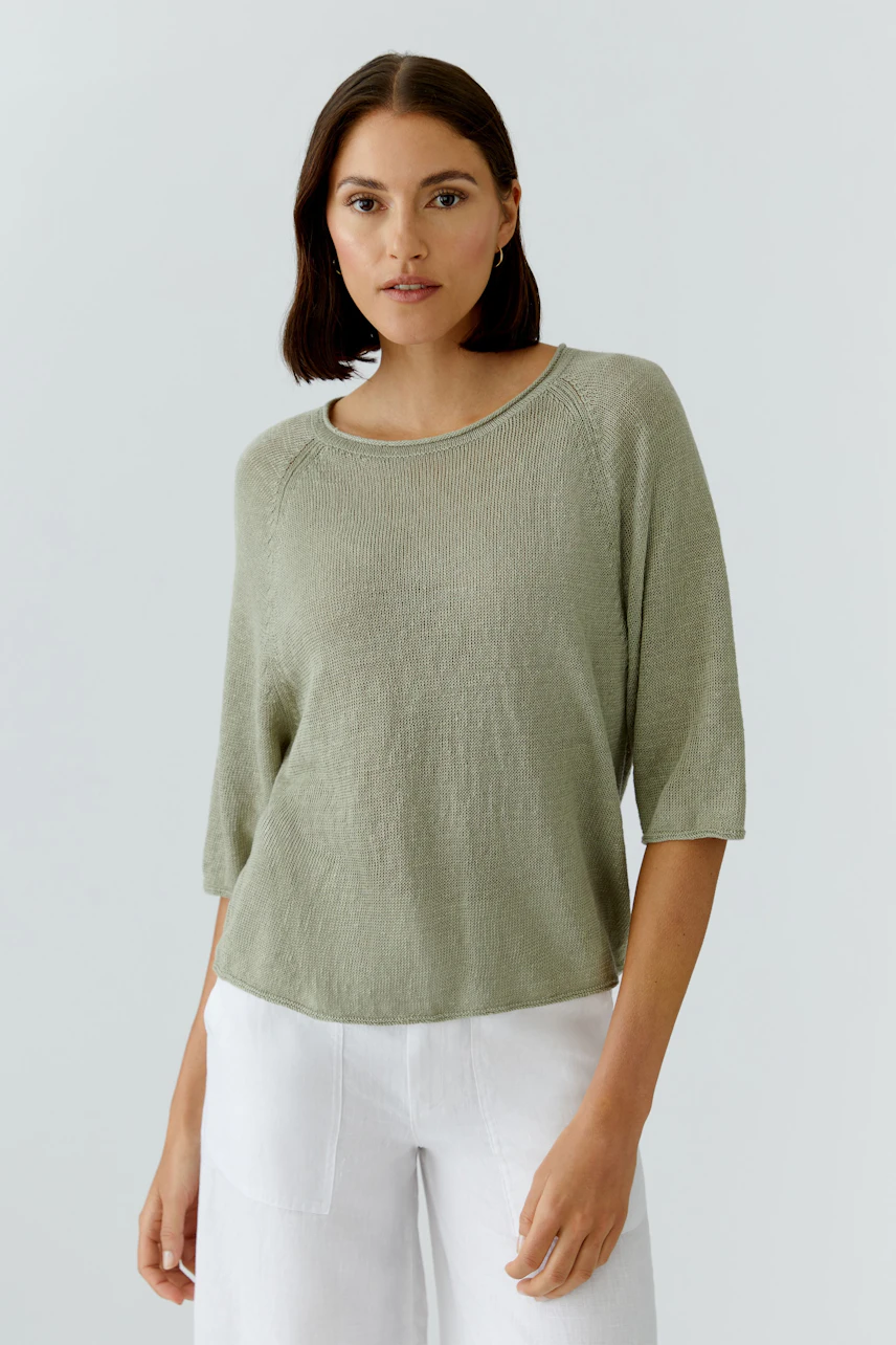 Lässiger Damen-Pullover: Frau im grünen Pullover mit halblangen Ärmeln und weißen Hosen schaut entspannt.