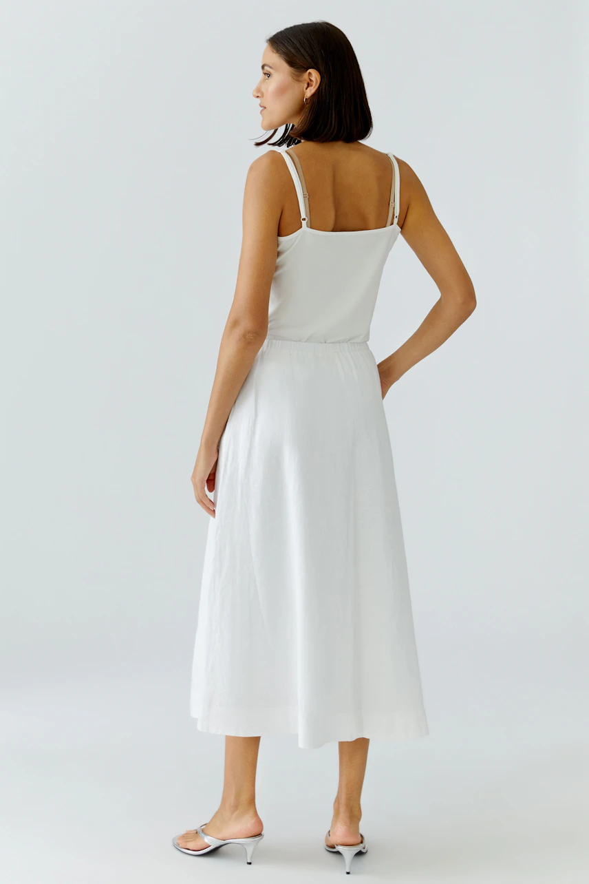 A-Linien-Rock: Frau in weißem, knielangem Rock und Top, seitlich stehend, mit schlichten Sandalen.