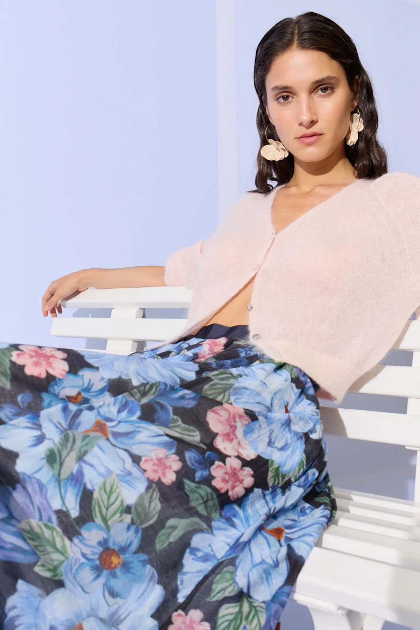 Woman in a soft pink cardigan with floral skirt, sitting confidently on a white bench.