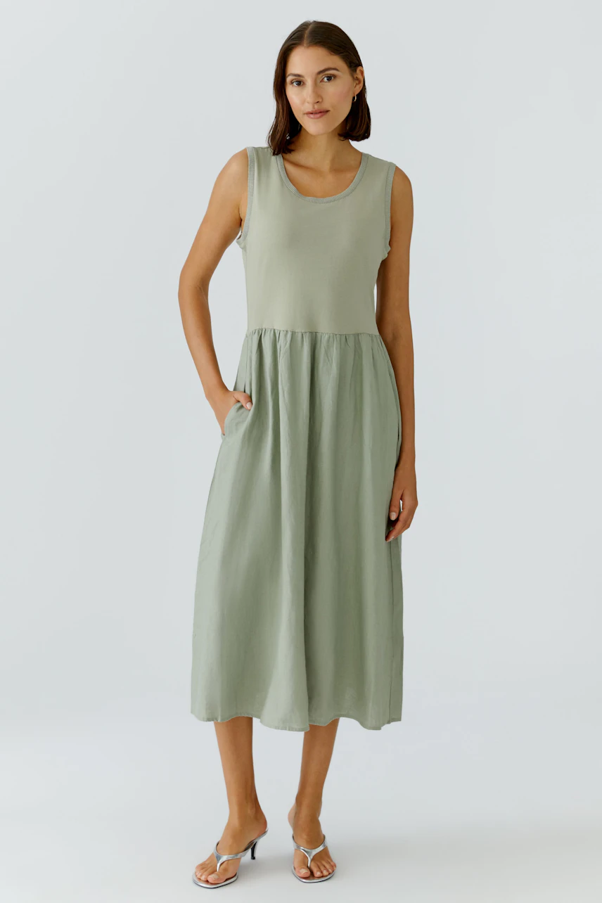 Casual midi dress: Woman in sleeveless green dress with relaxed fit and side pockets, standing confidently.