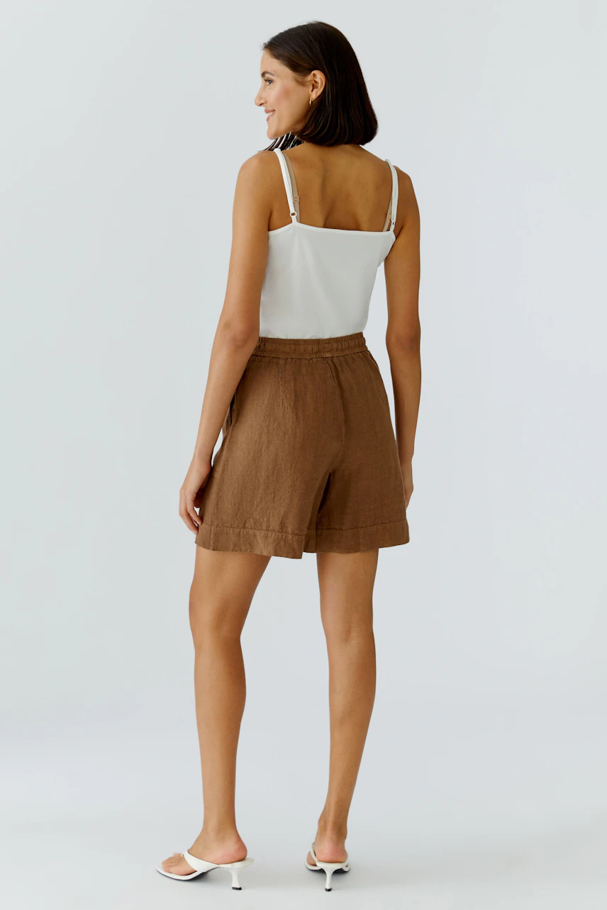Casual shorts: Woman in brown shorts and white top, standing confidently in white sandals.