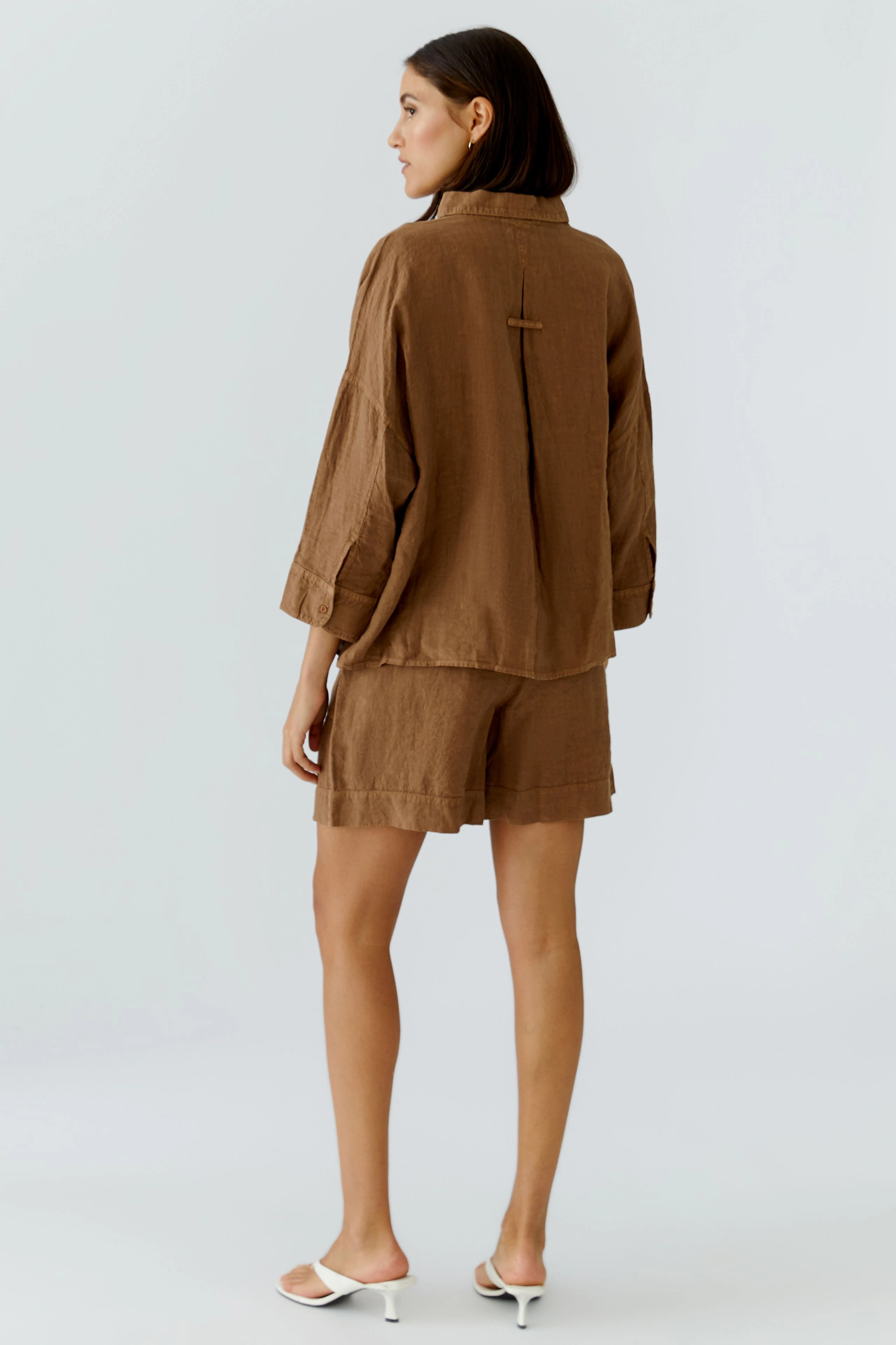 Lässige Damen-Bluse: Frau in brauner Bluse und Shorts, seitlich stehend, mit schlichten weißen Sandalen.