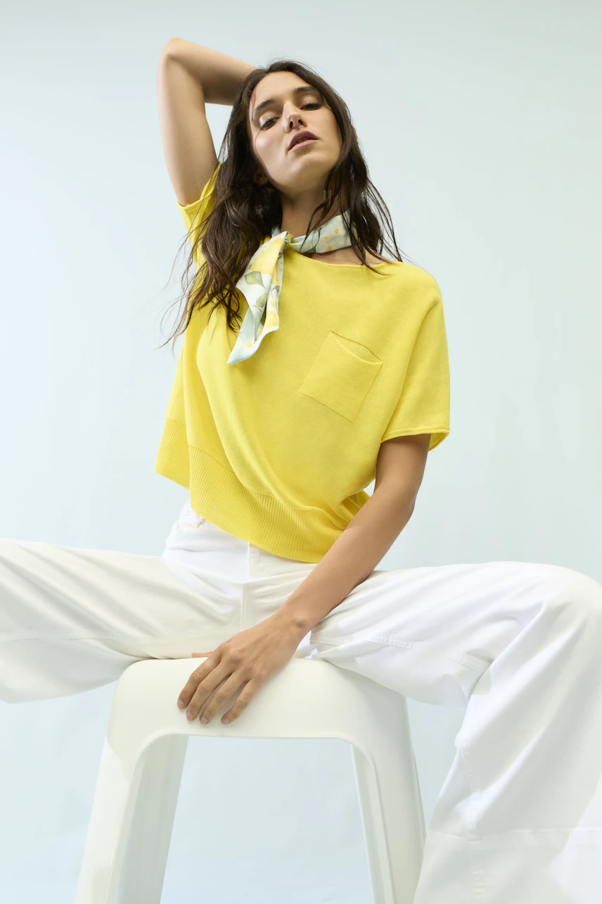 Woman in yellow top and white wide leg jeans, sitting confidently on a stool, with a patterned scarf.