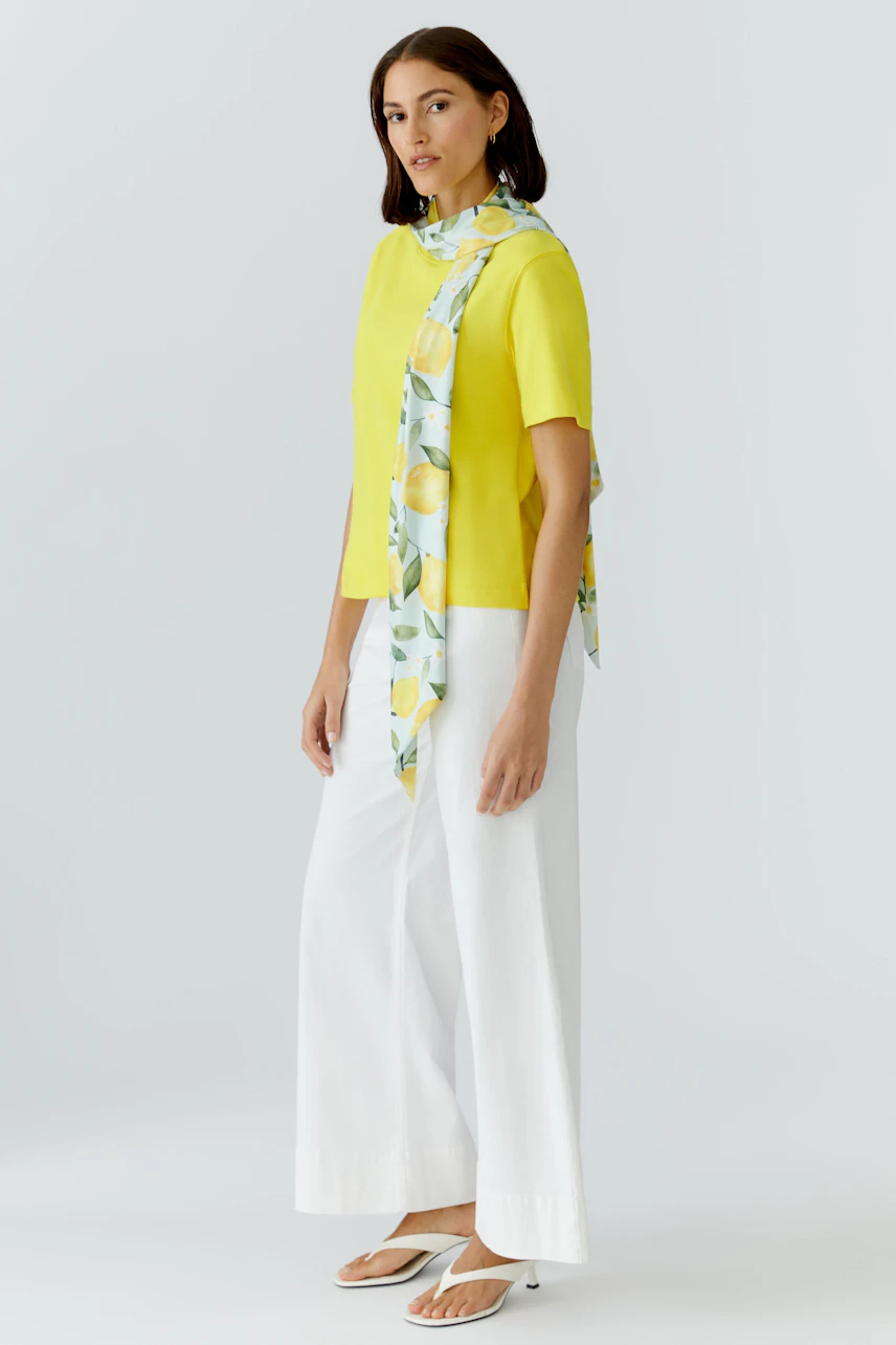 Woman in a lemon-patterned scarf, yellow top, and white pants, standing confidently.