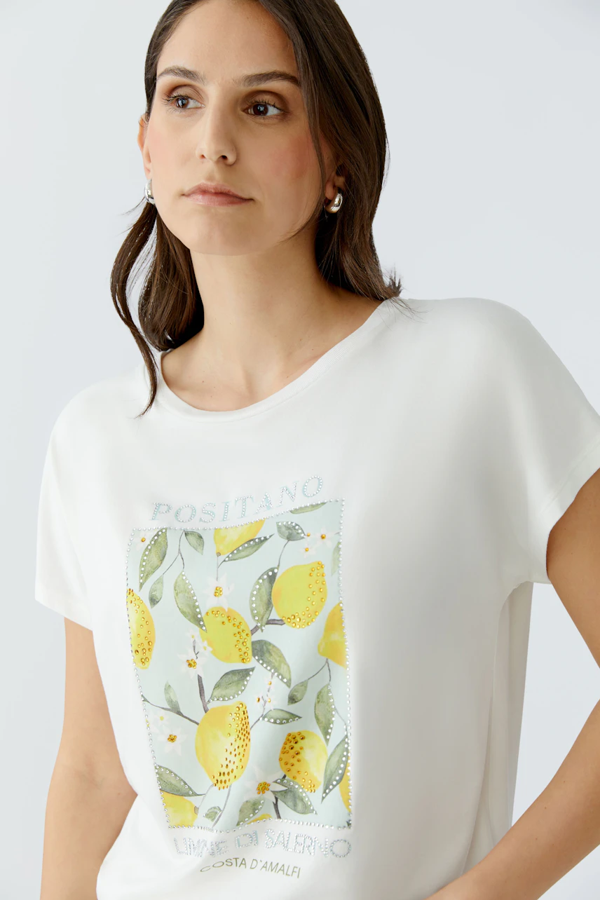 Damen-T-Shirt: Frau im weißen T-Shirt mit Zitronenprint und Schriftzug "Positano" blickt entspannt.