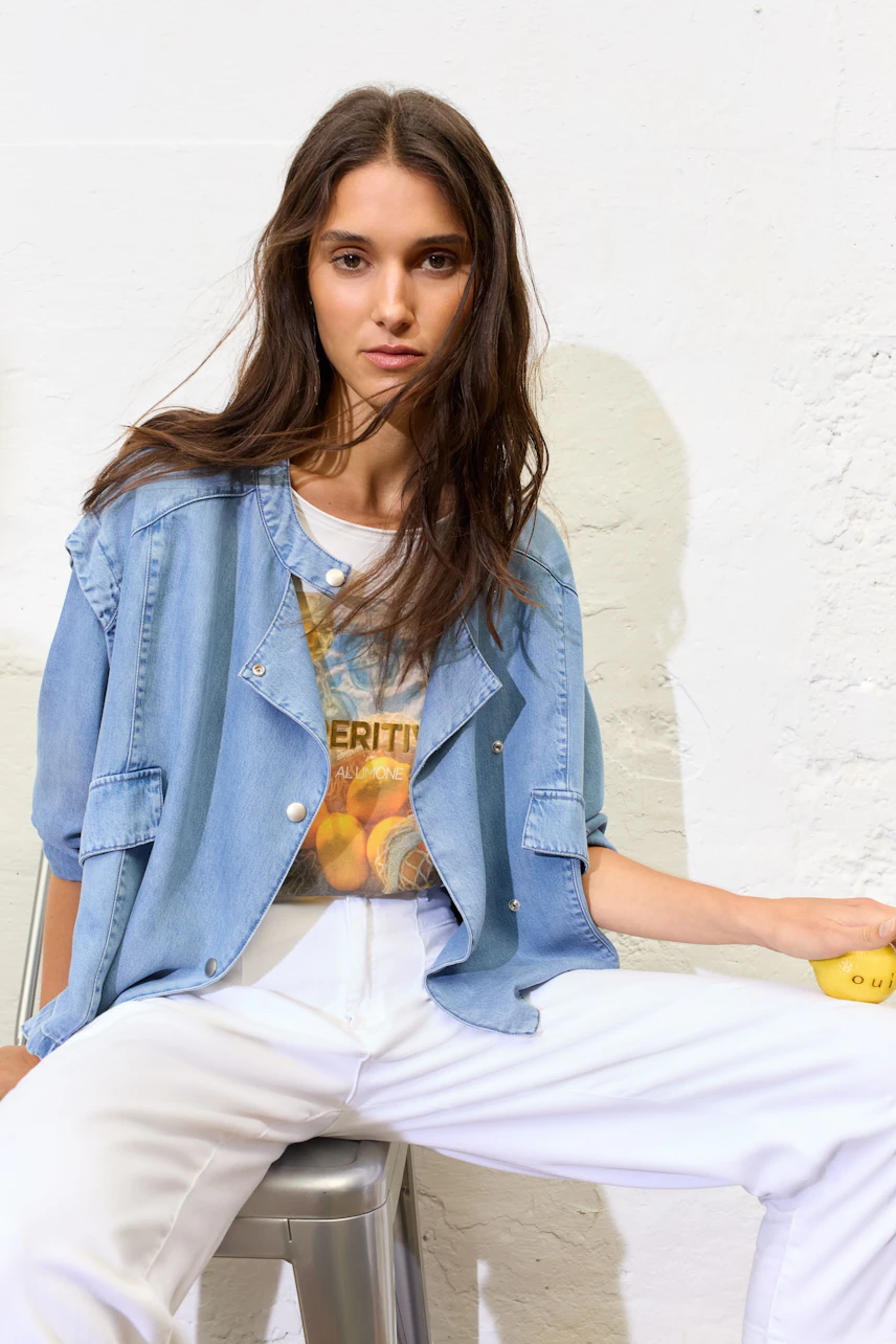 Lässiges Damen-T-Shirt: Frau im bedruckten T-Shirt mit Jeansjacke und weißen Hosen sitzt entspannt.