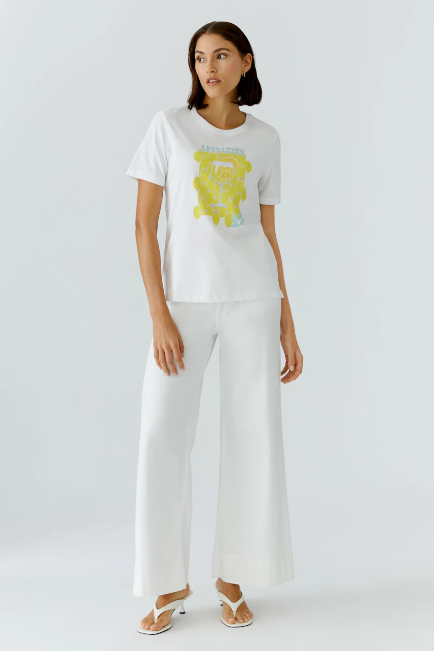 Weißes Damen-T-Shirt: Frau trägt T-Shirt mit gelbem Print, kombiniert mit weißer Hose und Sandalen.