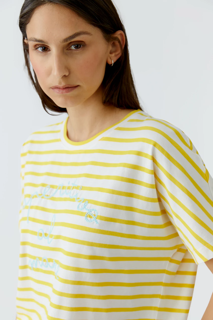 Gestreiftes Damen-T-Shirt: Frau im gelb-weiß gestreiften T-Shirt mit dezentem Schriftzug blickt ernst.