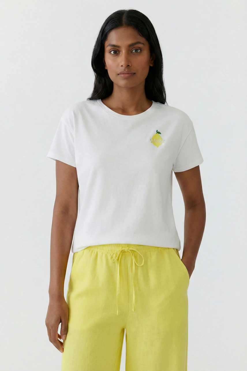 Weißes Damen-T-Shirt: Frau im T-Shirt mit Zitronenmotiv und gelber Hose, schlicht und sommerlich.