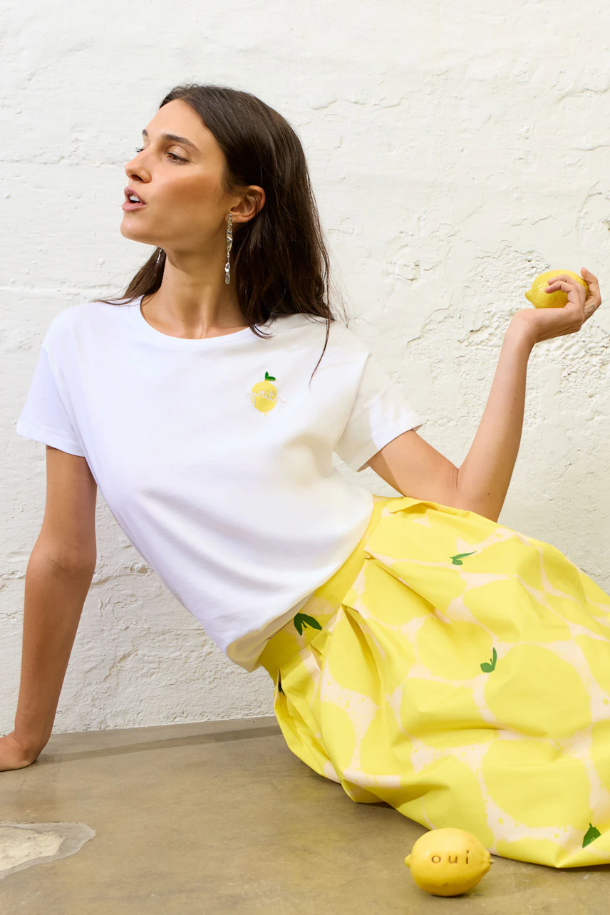 Weißes T-Shirt mit Zitronenmotiv: Frau trägt es mit gelbem Rock, hält Zitrone in der Hand, sitzt lässig.