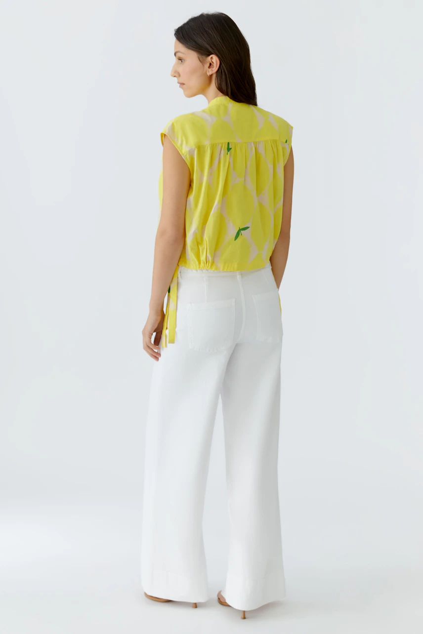 Frau in gelber Bluse mit Muster, kombiniert mit weißen Hosen. Rückansicht zeigt lässigen, sommerlichen Look.
