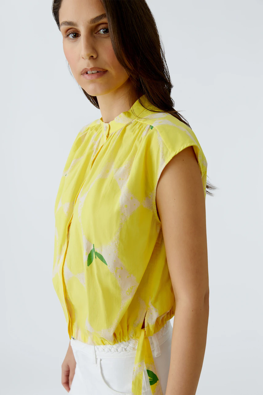 Frau in gelber Bluse mit Muster, ärmellos, kombiniert mit weißer Hose, schaut zur Seite.