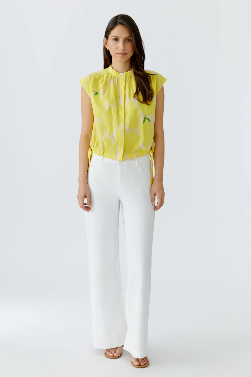 Frau in gelber Bluse mit Muster, kombiniert mit weißen Hosen. Frischer Look für den Sommer.