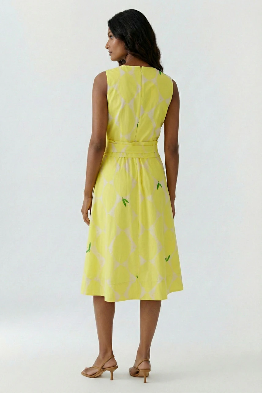 Frau im gelben Midikleid mit Muster, ärmellos, knielang, steht seitlich, trägt Sandalen.