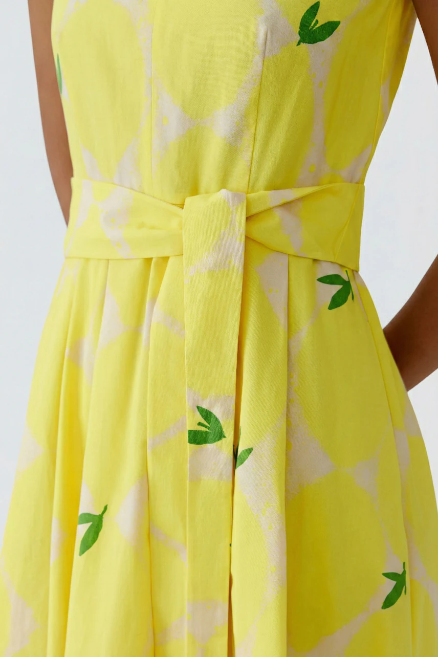 Gelbes Midikleid mit grünem Blattmuster und Bindegürtel, getragen von einer Frau.