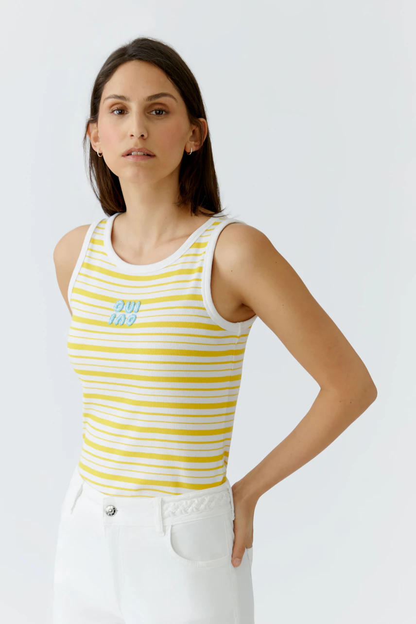 Junge Frau im gestreiften Tank Top in Gelb-Weiß, kombiniert mit weißen Hosen, schaut selbstbewusst.