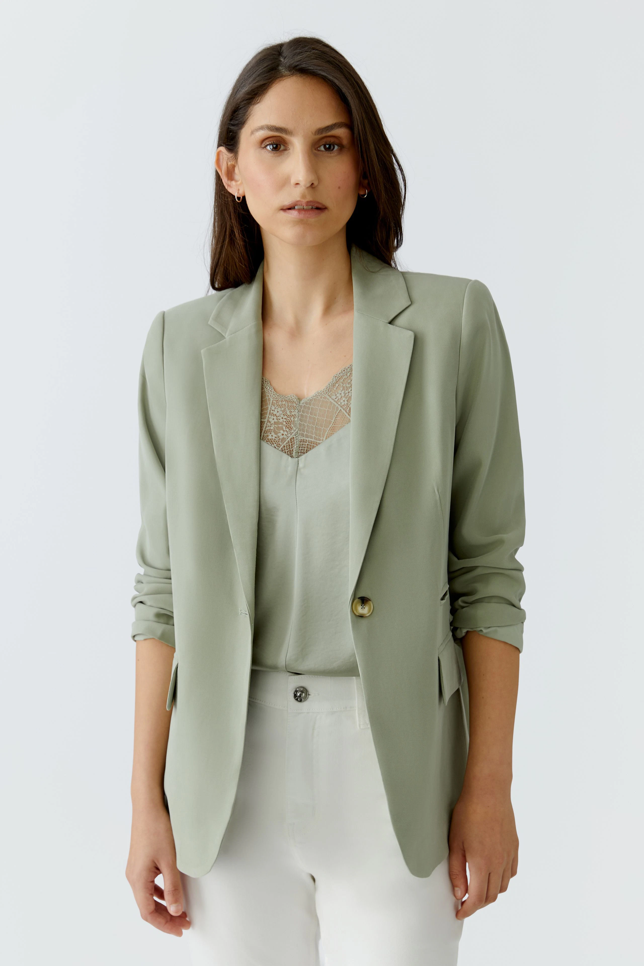 Casual women's blazer: Woman in light green blazer over a lace-trimmed top and white pants, looking poised.