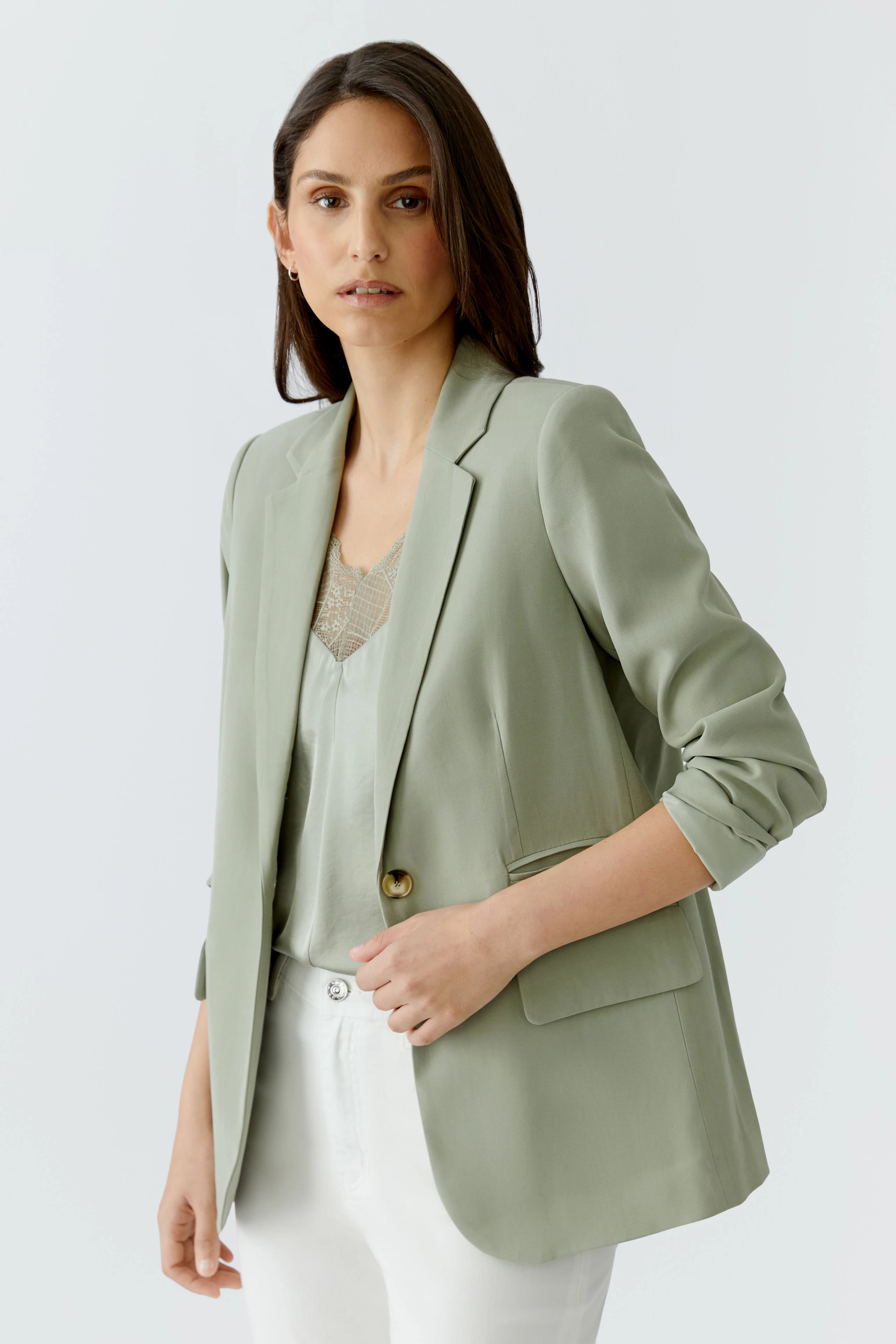 Casual women's blazer: Woman in light green blazer over a lace top and white pants, looking poised.