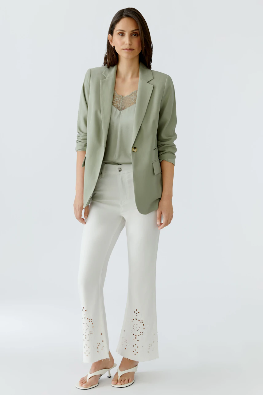 Casual women's blazer: Woman in a light green blazer over a lace top, paired with white pants and sandals.