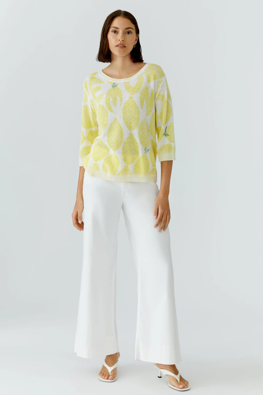 Frau im gelben Pullover mit Blattmuster, kombiniert mit weißen, weiten Hosen und Sandalen.