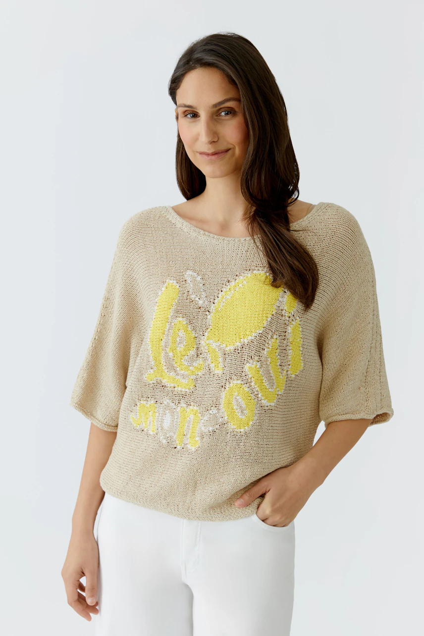 Lässiger Damen-Pullover: Frau im beigen Pullover mit gelbem Print und weißen Hosen lächelt entspannt.