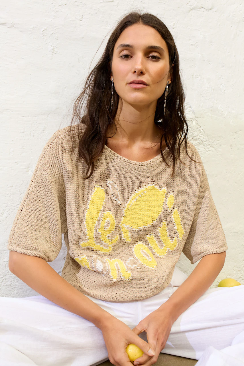 Lässiger Damen-Pullover: Frau im beige Pullover mit gelbem Print und weißen Hosen sitzt entspannt.