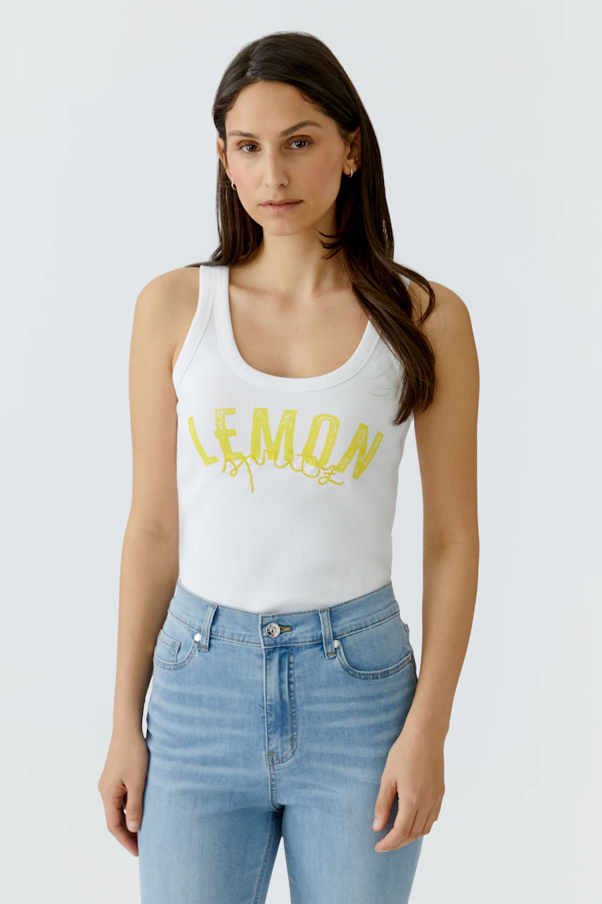 Junge Frau im weißen Tank Top mit gelbem Schriftzug, kombiniert mit hellblauer Jeans, schaut direkt.