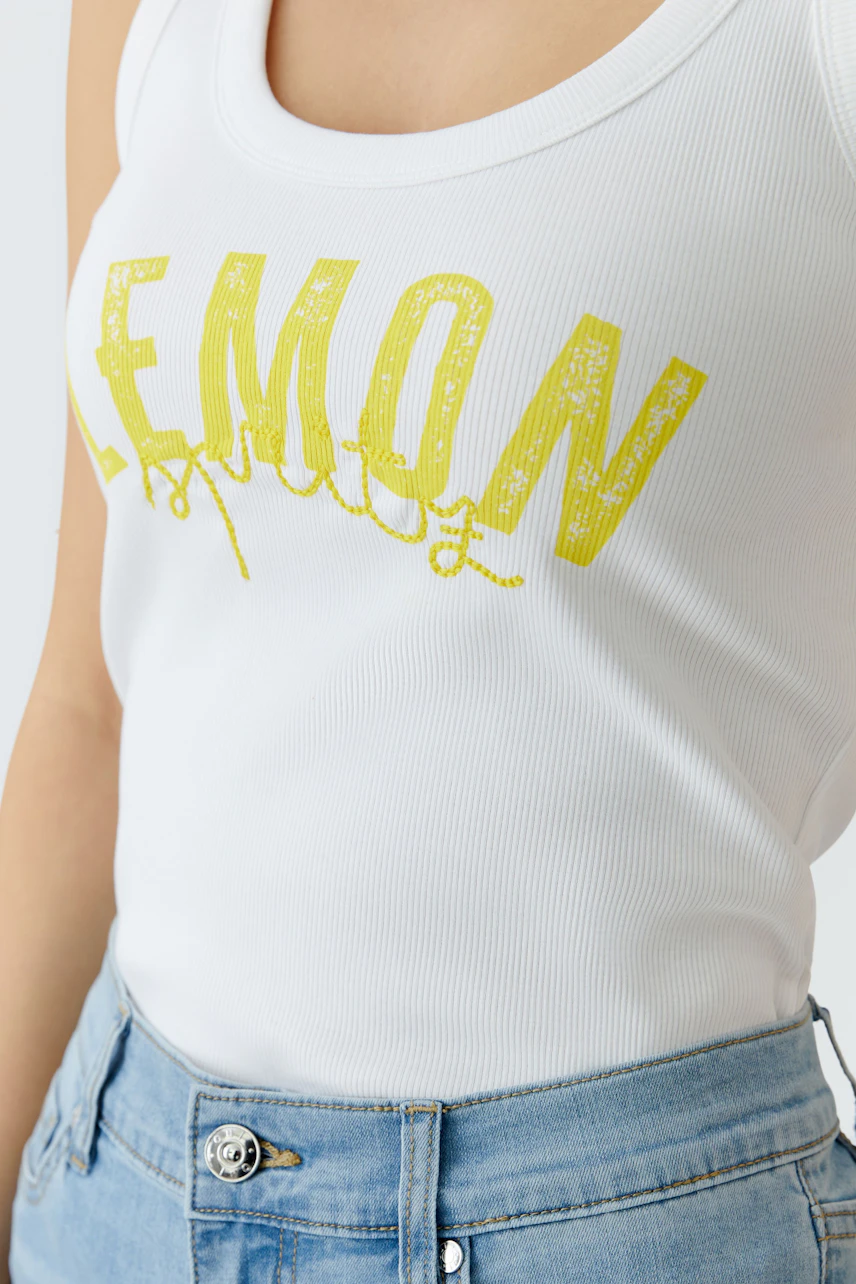Weißes Tank Top: Frau trägt ein ärmelloses Top mit gelbem Schriftzug, kombiniert mit blauer Jeans.