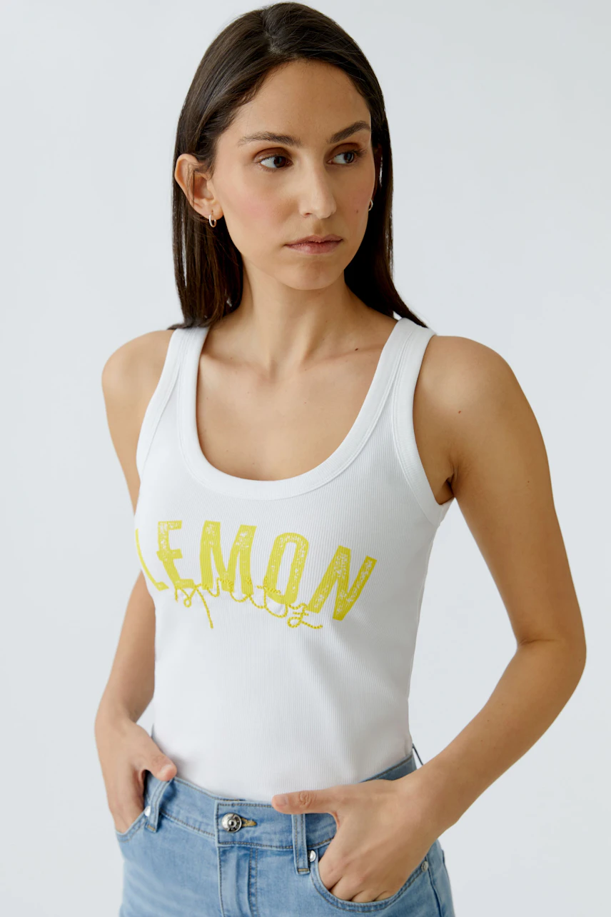Weißes Tank Top: Frau im lässigen Top mit gelbem "LEMON"-Print, kombiniert mit blauen Jeans.