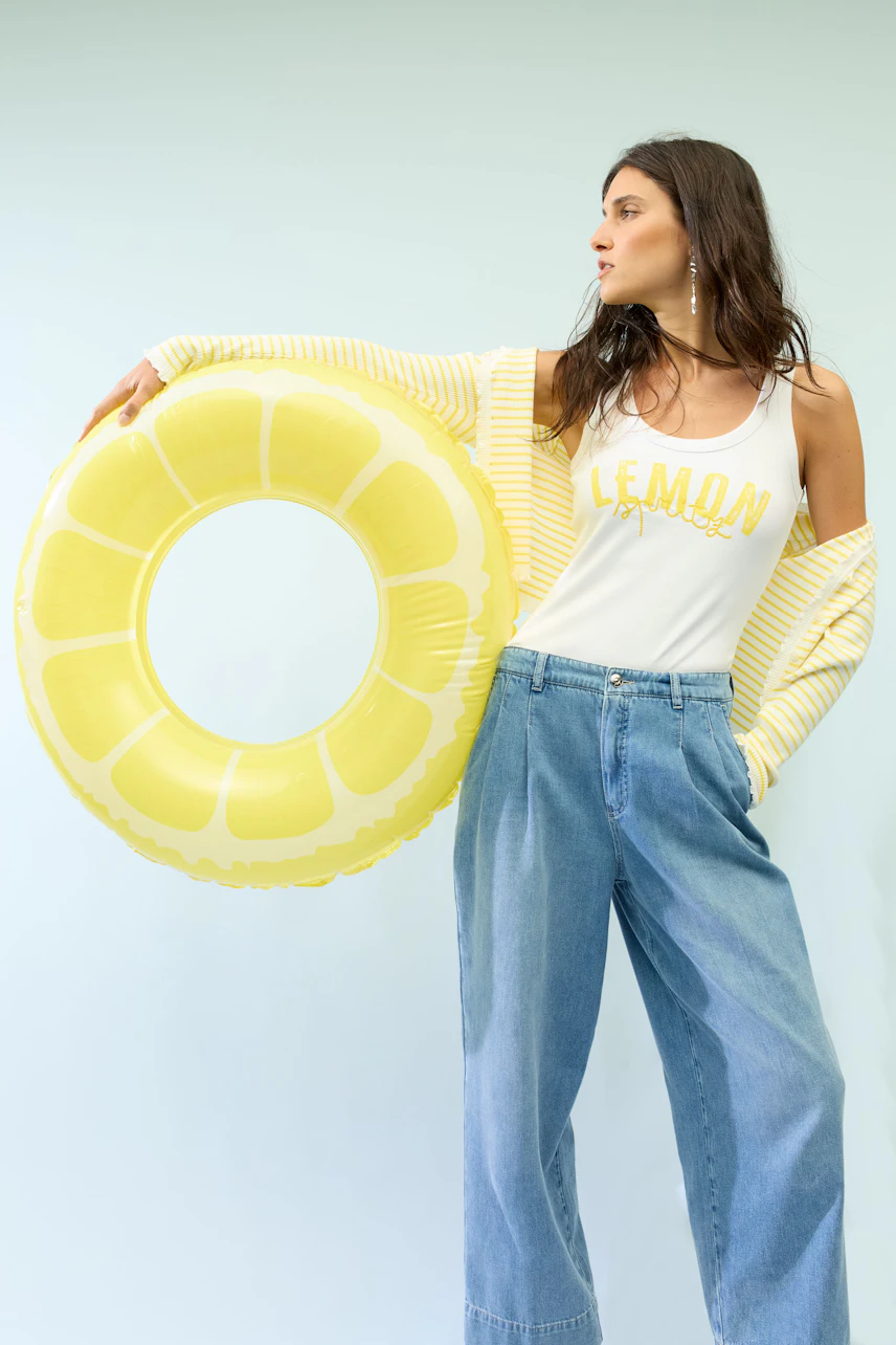 Junge Frau im weißen Tank Top mit "Lemon"-Aufdruck, gelber Jacke und blauer Jeans, hält gelben Schwimmreifen.