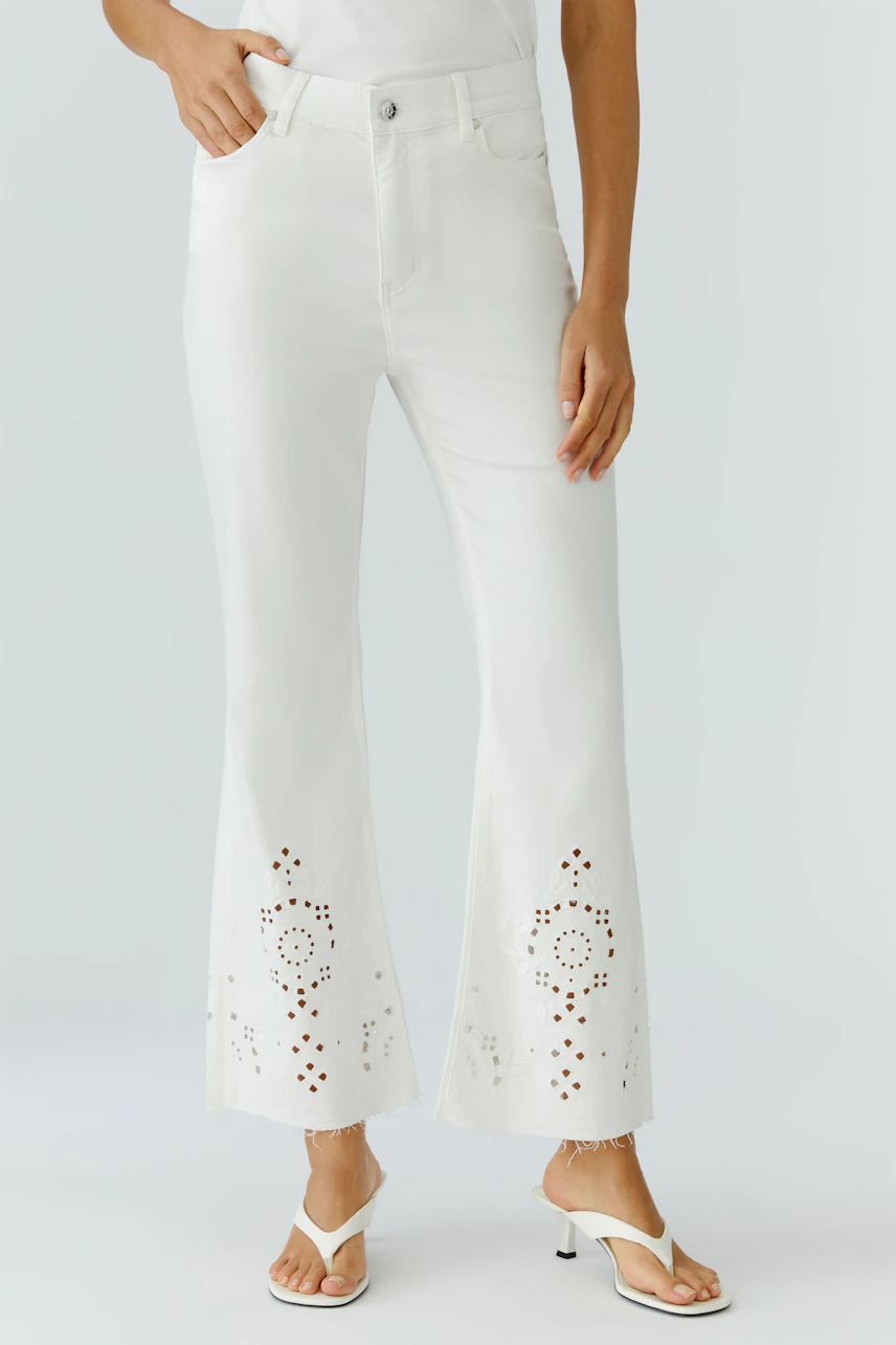 Frau in weißer Bootcut-Jeans mit Lochmuster, kombiniert mit weißen Sandalen. Lässig und modern.