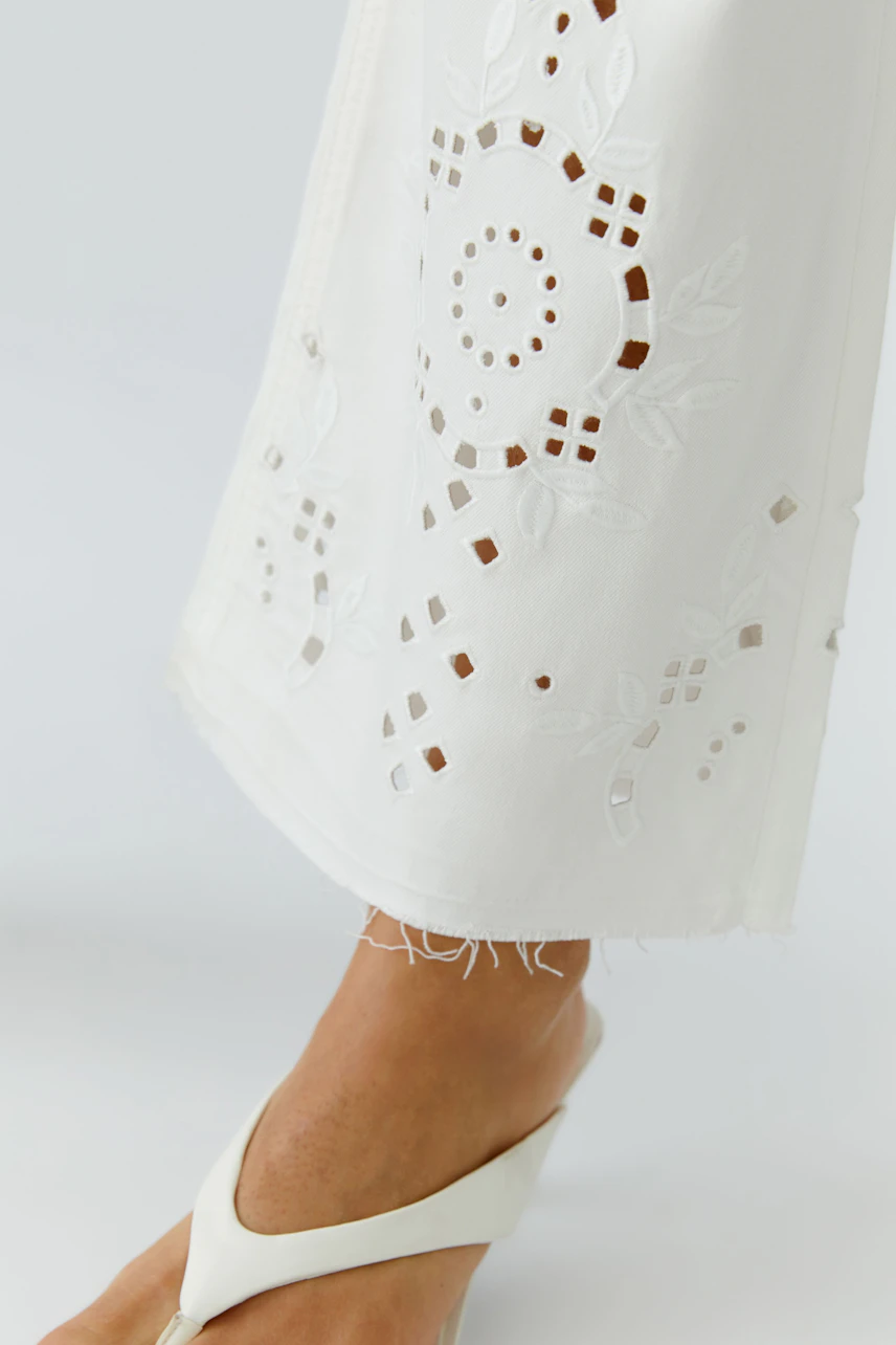 Weiße Bootcut-Jeans mit Lochstickerei, getragen von einer Frau in weißen Sandalen.