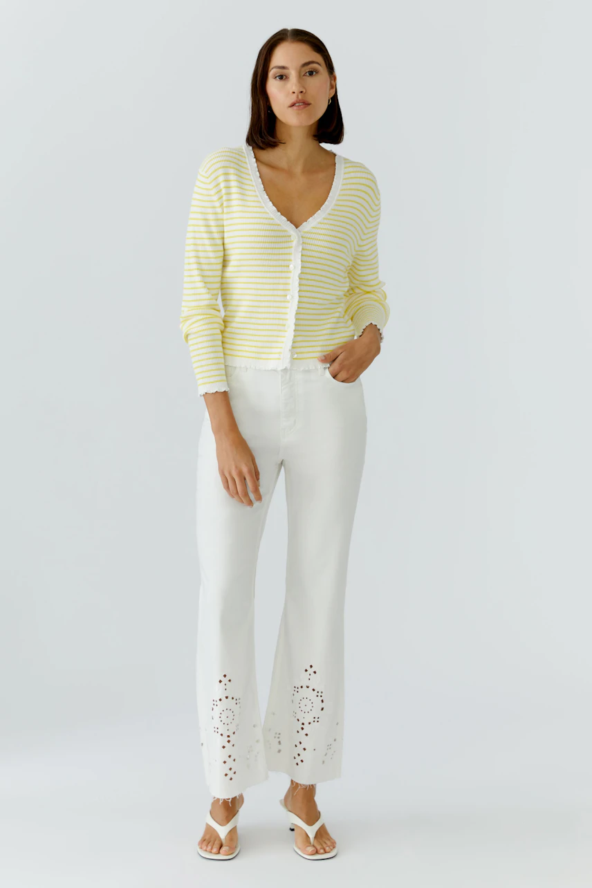 Frau in weißer Bootcut-Jeans mit Lochmuster, gelb-weiß gestreiftem Cardigan und Sandalen.