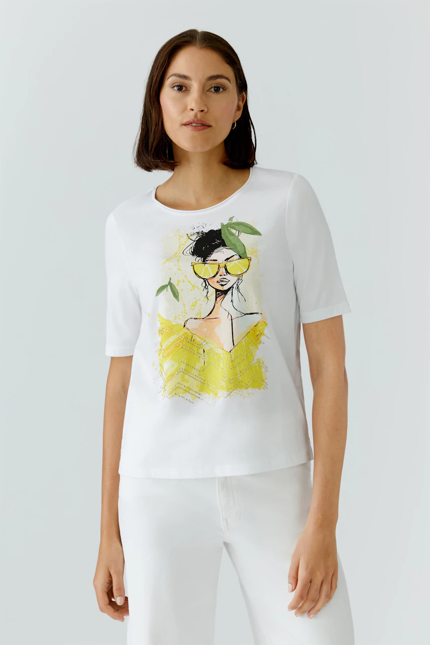 Damen-T-Shirt: Frau im weißen T-Shirt mit gelbem Grafikdruck und kurzen Ärmeln, kombiniert mit weißer Hose.