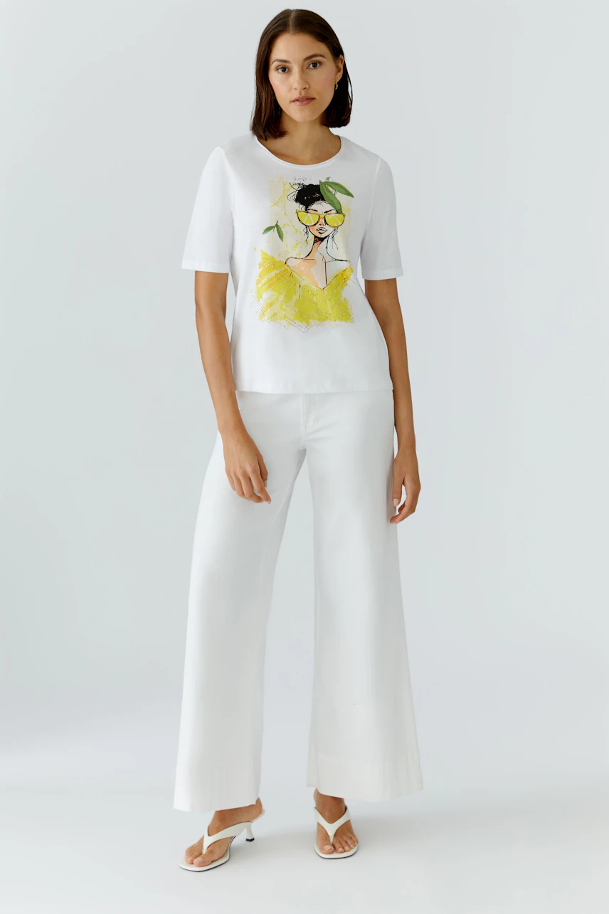 Weißes Damen-T-Shirt: Frau trägt T-Shirt mit grafischem Print und weiten weißen Hosen, steht selbstbewusst.
