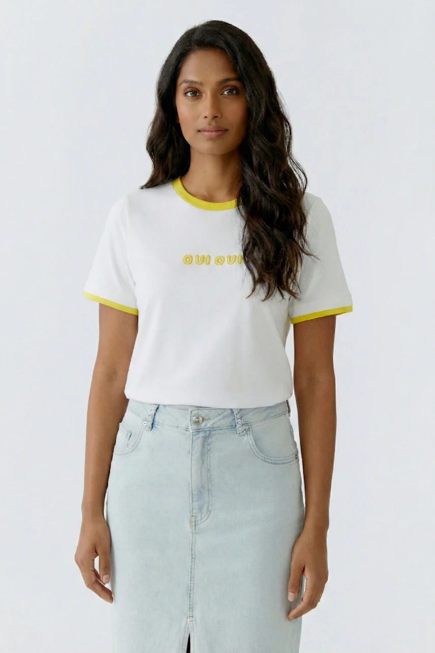 Weißes Damen-T-Shirt: Frau im T-Shirt mit gelben Details und heller Jeans blickt direkt in die Kamera.