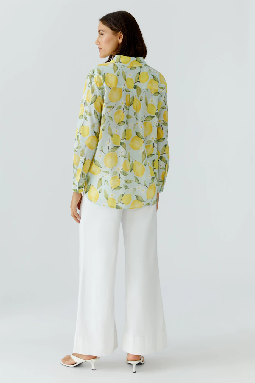 Frau in Hemdbluse mit Zitronenmuster, kombiniert mit weißen Hosen und Sandalen, blickt zur Seite.