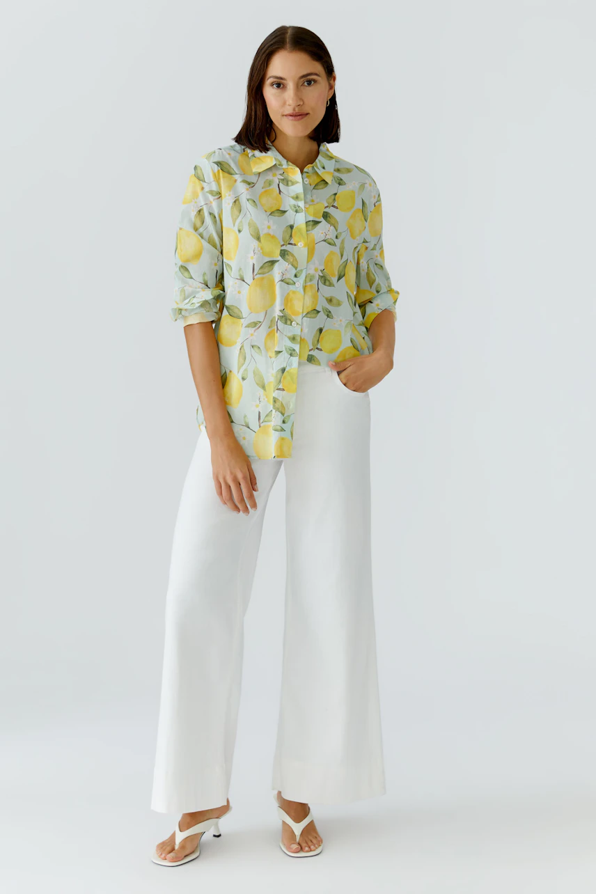 Frau in Hemdbluse mit Zitronenmuster, kombiniert mit weißen Hosen und Sandalen, lässig und frisch.