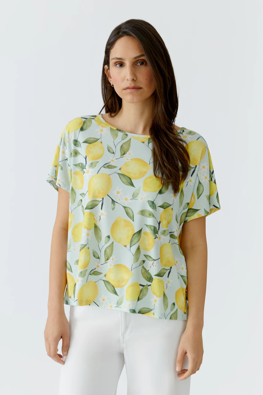 Damen-T-Shirt: Frau im hellen T-Shirt mit Zitronenmuster und weißen Hosen, lässiger Look.