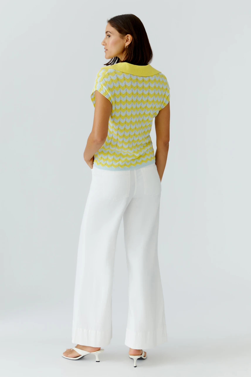 Frau im gelben Pullover mit Zickzack-Muster, kombiniert mit weißen Hosen und Sandalen, von hinten gesehen.