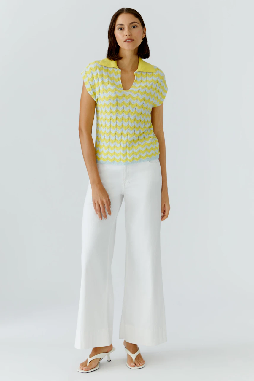 Frau im gelben Pullover mit Muster, kombiniert mit weißen Hosen und Sandalen, steht selbstbewusst.
