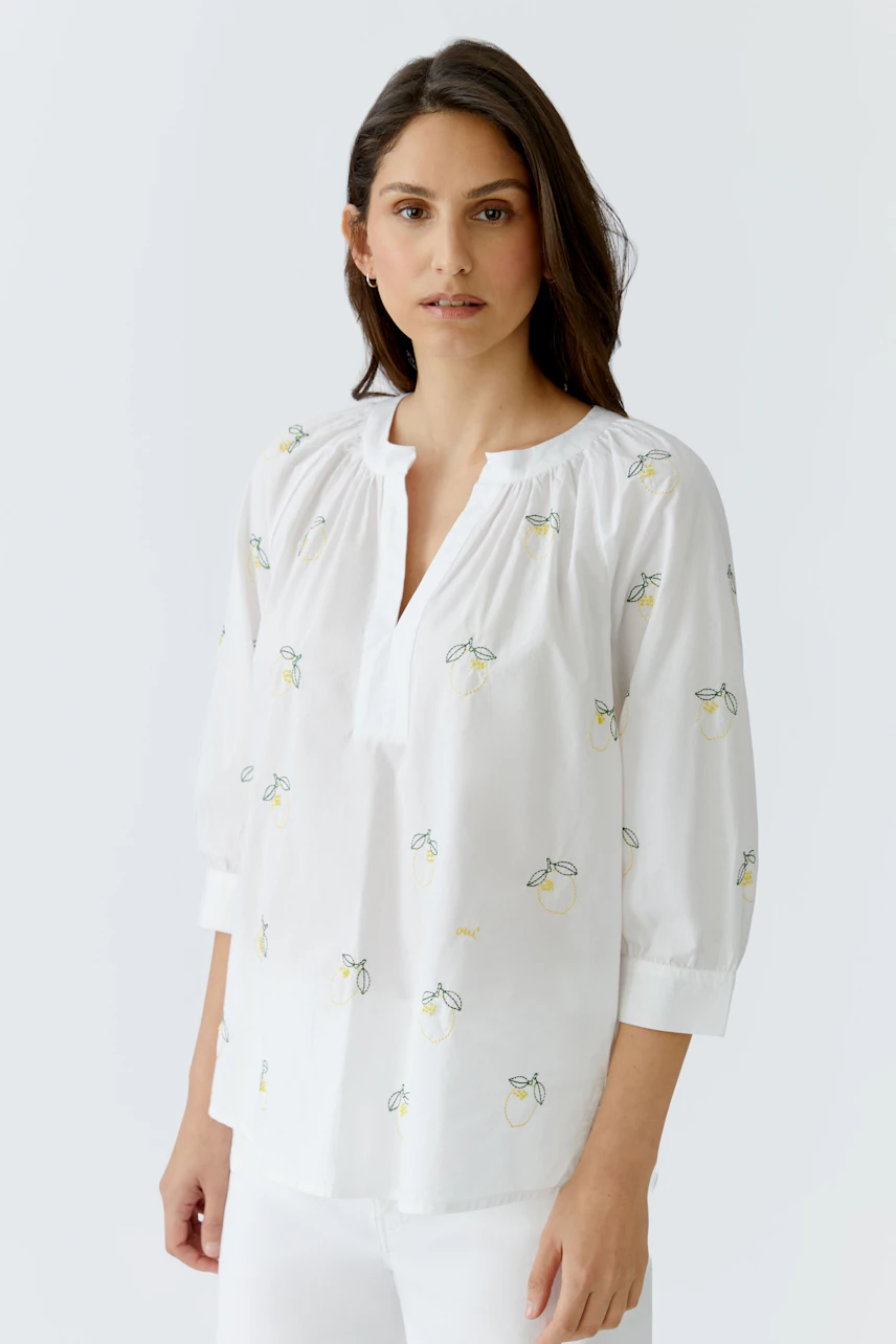 Weiße Tunika: Frau in lockerer Tunika mit Zitronenmotiv und Dreiviertelärmeln, kombiniert mit weißer Hose.