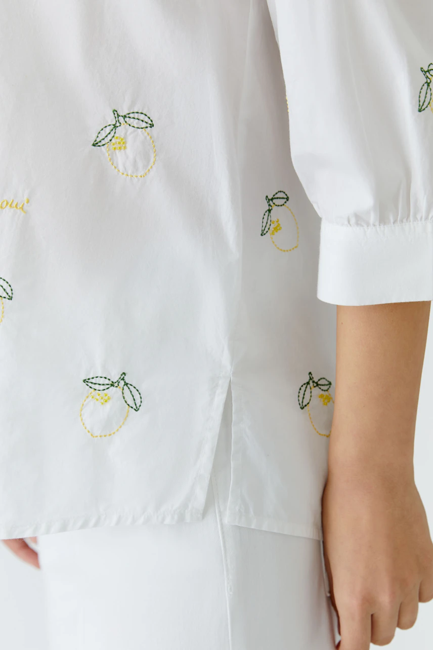Weiße Tunika mit Zitronenstickerei: Frau trägt lässige Tunika mit 3/4-Ärmeln, kombiniert mit weißer Hose.